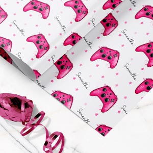 May include: Wrapping paper featuring a repeating pattern of bright pink video game controllers and small pink stars on a white background. The name Samantha is printed in a black cursive script throughout the design. A pink ribbon is included.