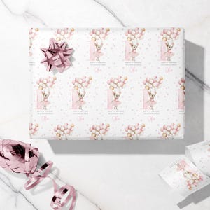 May include: White wrapping paper with a repeating pattern of a ballerina holding balloons, the number 1, and the name "Nora". A pink bow and ribbon are on the marble surface. The text reads "Happy 1st Birthday to a special niece."