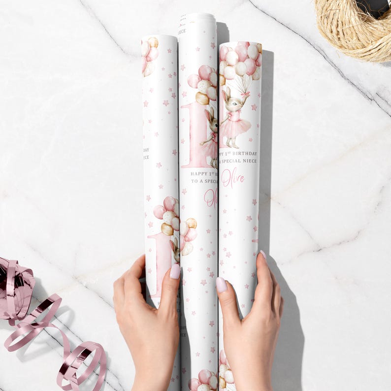 May include: Three rolls of white wrapping paper with a pink and gold design. The paper features a bunny holding balloons, the number 1, and the text "Happy 1st Birthday to a Special Niece". Pink stars are scattered across the paper.