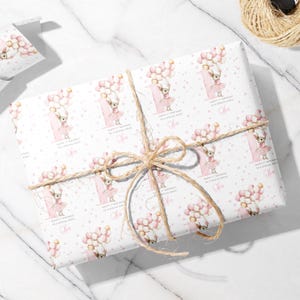 May include: White wrapping paper with a repeating pattern of a cartoon chihuahua wearing a pink tutu and holding balloons. The paper is tied with twine and features text that reads "Happy 1st Birthday to a Special Niece" and a name.