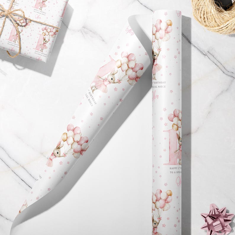 May include: White wrapping paper with a repeating pattern of a bunny holding balloons, pink stars, and the number 1. The text reads "Happy 1st Birthday to a Special Niece". A wrapped gift and a pink bow are also visible.