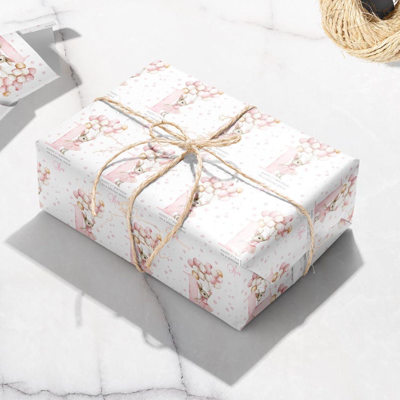 May include: A wrapped gift box with white wrapping paper featuring a repeating pattern of a bunny holding balloons in pink and peach colors. The box is tied with twine.
