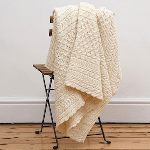 Könnte beinhalten: Eine cremefarbene Strickdecke mit Zopf- und strukturierten Mustern, die über einem kleinen Holzstuhl drapiert ist. Die Decke hat einen gewellten Rand und besteht aus einem weichen, warmen Material, ideal, um jedem Raum eine gemütliche Note zu verleihen.