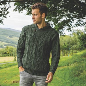 Könnte beinhalten: Ein dunkelgrüner Zopfstrickpullover mit Schalkragen und einem einzelnen Holzknopfverschluss. Der Pullover hat aufwendige Webmuster und lange Ärmel. Die Person trägt eine graue Jeans.