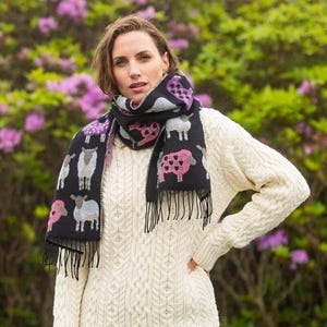 Könnte beinhalten: Schwarzer Schal mit Schafmuster in Rosa, Lila und Grau. Der Schal hat Fransen und wird über einem cremefarbenen Zopfstrickpullover getragen. Der Hintergrund zeigt grünes Laub und rosa Blüten.