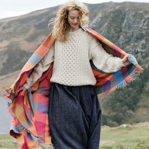 May include: A person wearing a cream cable-knit jumper and a dark blue skirt, with a colourful plaid blanket draped over them. The blanket has orange, pink, blue, and green squares with fringed edges. The background shows a mountain landscape.