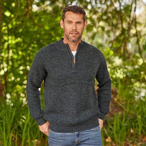 May include: A dark gray, textured knit sweater with a quarter-zip closure and a brown leather pull. The sweater has a ribbed design and long sleeves. The person is wearing blue jeans.