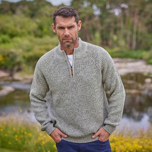May include: A light green zip-up sweater with a ribbed texture. The sweater has a high collar and a brown zipper pull. The person is wearing blue jeans. The background is a natural outdoor setting with trees and water.