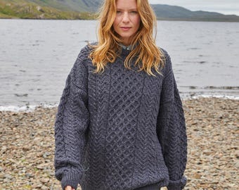 May include: A dark gray, cable-knit sweater with a textured diamond pattern. The sweater has a crew neck and long sleeves with ribbed cuffs. The model is standing on a rocky beach with a lake and mountains in the background.