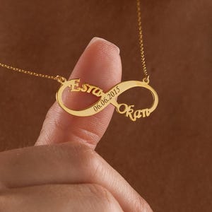 Peut inclure: Un collier infini doré avec les noms "Esto" et "Okan" gravés de chaque côté du symbole infini. La date "06.06.2015" est également gravée. Le collier est présenté par un doigt, mettant en valeur le design.