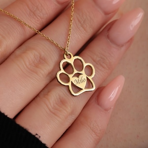 May include: A gold-colored necklace featuring a paw print pendant with a heart-shaped cutout. The word "Milo" is engraved on the heart. The necklace is displayed on a person's hand.