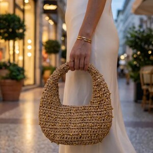 Könnte beinhalten: Eine halbmondförmige Handtasche aus geflochtenem beigefarbenem Strohmaterial mit einem abgerundeten Tragegriff, verziert mit kleinen, gleichmäßig verteilten goldfarbenen Metallnieten auf der Außenfläche, die für eine strukturierte Optik sorgen.