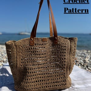 May include: A brown crochet tote bag with leather handles. The bag has a woven design with a rectangular shape. The text "Crochet Pattern" is displayed in black. The bag is on a beach with the ocean in the background.