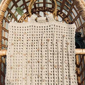 Puede incluir: Bolso tote de macramé color crema con asa de bambú. El bolso presenta un diseño tejido con pequeños detalles decorativos y flecos en la parte inferior. El bolso se exhibe en una silla de mimbre.