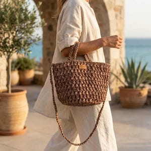 May include: A brown woven tote bag featuring a textured, braided design with two short handles and a long, matching braided shoulder strap. A small rectangular label is attached to the front center of the bag, displaying visible text.
