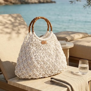 May include: A white macrame handbag featuring a knotted cord design and two circular bamboo handles. A small rectangular leather-coloured label is attached to the front centre, displaying the text "Sea Design Studio" in a dark, clear font.