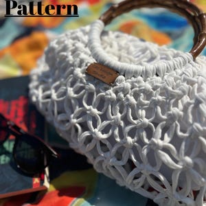 May include: White crochet bag with a bamboo handle. The bag has a knotted design and a small leather tag that reads "Car design Studio". The words "Crochet Pattern" are at the top of the image.