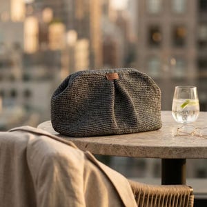 May include: A dark gray, textured, woven clutch bag with a rounded, pleated silhouette and a small, rectangular brown leather-like brand tag centered on the top edge. The bag features a firm, structured frame and a dense, consistent knit pattern.