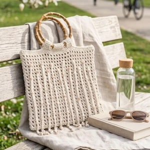 May include: A cream-colored crocheted tote bag featuring vertical fringe detailing and two circular bamboo handles. The bag is accompanied by a clear glass water bottle with a cork stopper, a closed book, and beige-framed sunglasses.