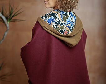 May include: A burgundy cape with a tan-colored hood and a patterned lining featuring blue, green, and white floral designs. The cape is made of a soft material and is worn over black clothing.