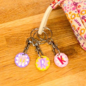 May include: Three handmade keychains with floral designs and the letters "M", "R", and "K". Each keychain has a silver clasp and is attached to a keyring. A floral patterned bag is in the background.