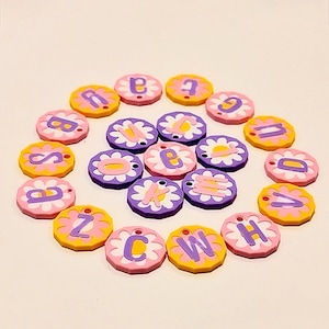 May include: A collection of colorful, round charms with letters of the alphabet. Each charm has a flower-shaped border in pink, yellow, or purple, with a contrasting letter in the center. The charms are arranged in a circular pattern.
