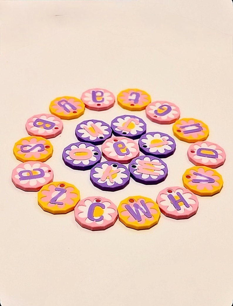 May include: A collection of colorful, round charms with letters of the alphabet. Each charm has a flower-shaped border in pink, yellow, or purple, with a contrasting letter in the center. The charms are arranged in a circular pattern.