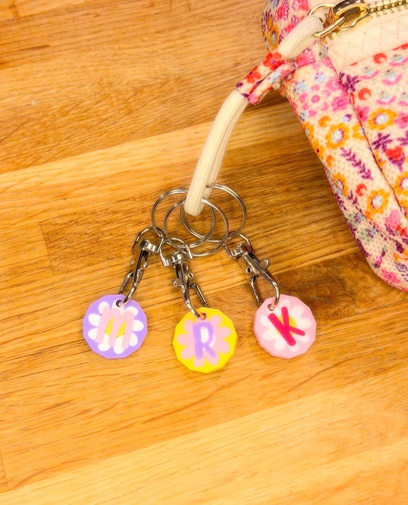 May include: Three handmade keychains with floral designs and the letters "M", "R", and "K". Each keychain has a silver clasp and is attached to a keyring. A floral patterned bag is in the background.