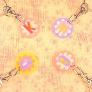 May include: Four flower-shaped keychains with silver clasps. Each keychain has a different color flower with a letter in the center. The flowers are pink, yellow, and purple. The letters are K, M, R, and B.