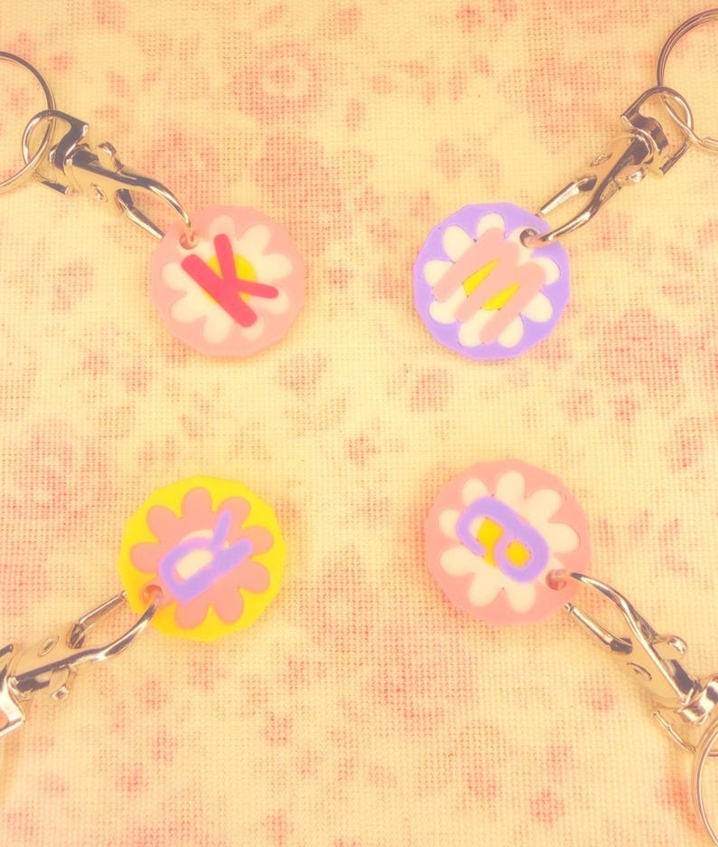 May include: Four flower-shaped keychains with silver clasps. Each keychain has a different color flower with a letter in the center. The flowers are pink, yellow, and purple. The letters are K, M, R, and B.
