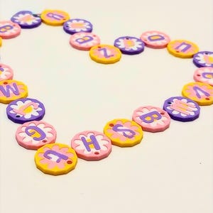 May include: A heart shape made of colorful flower-shaped beads. Each bead has a letter in the center. The beads are pink, yellow, and purple, with white flower petals. The letters are in a contrasting color.