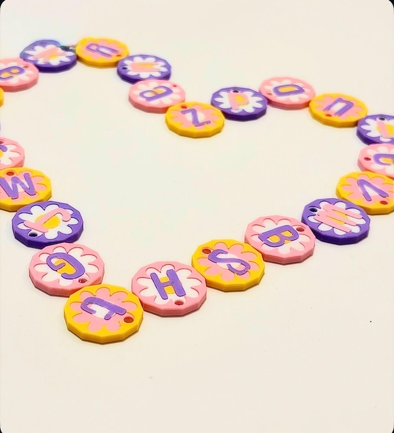 May include: A heart shape made of colorful flower-shaped beads. Each bead has a letter in the center. The beads are pink, yellow, and purple, with white flower petals. The letters are in a contrasting color.