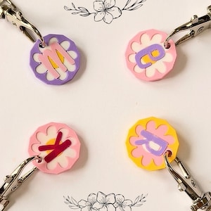 May include: Four flower-shaped keychains with silver clasps. Each keychain features a different letter: M, B, K, and R. The flowers have various color combinations, including pink, purple, and yellow, with white accents. Decorative floral illustrations are in the background.