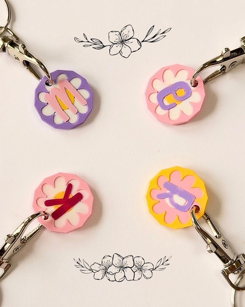 May include: Four flower-shaped keychains with silver clasps. Each keychain features a different letter: M, B, K, and R. The flowers have various color combinations, including pink, purple, and yellow, with white accents. Decorative floral illustrations are in the background.