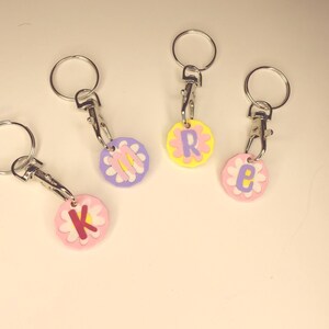 May include: Four keychains with flower-shaped charms. Each charm has a different letter: K, R, E, and M. The charms are in pastel colors: pink, yellow, and purple. Each keychain has a silver-colored ring and clasp.