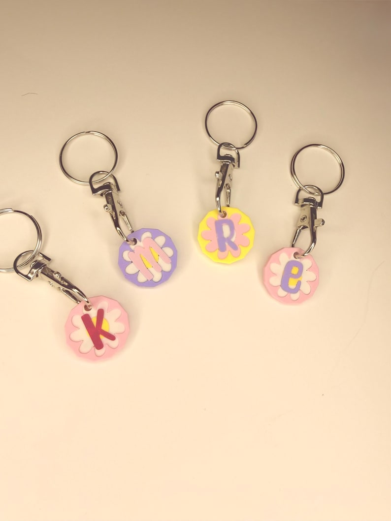 May include: Four keychains with flower-shaped charms. Each charm has a different letter: K, R, E, and M. The charms are in pastel colors: pink, yellow, and purple. Each keychain has a silver-colored ring and clasp.