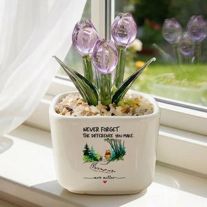 Könnte beinhalten: Ein weißer Keramiktopf mit einem Glas-Tulpenarrangement. Der Topf trägt den Text "NEVER FORGET THE DIFFERENCE YOU MAKE" und eine kleine Illustration. Die Tulpen sind lila und die Blätter sind grün.