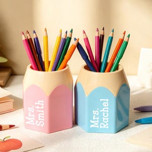 Puede incluir: Dos portalápices hexagonales con forma de lápiz, uno rosa y otro azul claro, cada uno con una sección superior beige. El portalápices rosa está personalizado con el texto blanco Mrs. Smith y el azul claro con el texto blanco Mrs. Rachel.