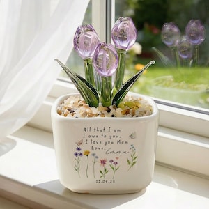 May include: A white square ceramic pot with a floral design and the text "All that I am I owe to you. I love you Mom Love, Emma 11.04.25." The pot contains decorative stones and glass tulip flowers with purple petals and green leaves.