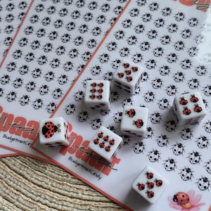 Può includere: Dadi bianchi con disegni di coccinelle rosse e pois neri sono sparsi su uno sfondo a tema coccinella. Lo sfondo presenta motivi di coccinelle e il testo "100-cent" in rosa. I dadi sono per un gioco.