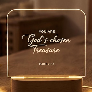 Può includere: Una luce notturna rettangolare in acrilico con angoli superiori arrotondati, inserita in una base di legno liscia. Il pannello trasparente è illuminato da una luce calda e riporta il testo bianco: YOU ARE God's chosen Treasure ISAIAH 41:10. Un cavo.