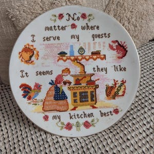 Könnte beinhalten: Ein weißer Zierteller mit goldenem Rand und Kreuzstichmuster. Das Design zeigt eine Küchenszene mit dem Text "No matter where I serve my guests, it seems they like my kitchen best."