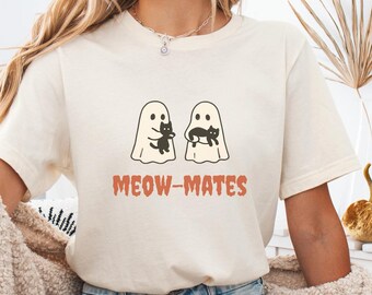 May include: Cream-colored t-shirt featuring a graphic of two ghost figures, each holding a black cat. The text "MEOW-MATES" is printed in a rust-colored font below the ghosts. The shirt is paired with denim shorts.