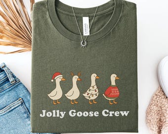 May include: Olive green t-shirt featuring a graphic of four geese dressed in holiday attire. The geese are wearing a Santa hat, reindeer antlers, a Christmas tree dress, and a red sweater. The text "Jolly Goose Crew" is below the geese.