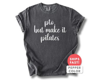 Op de afbeelding: Donkergrijs t-shirt met de witte tekst "pto, but make it pilates". Het shirt heeft opgerolde mouwen en een ronde hals. Een rood-witte sticker rechtsonder zegt "SHIPS FAST! PEPPER COLOR".