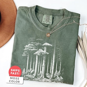 Op de afbeelding: Een mosgroen t-shirt met een bosafbeelding. Het shirt heeft een ronde hals en korte mouwen. Een gouden ketting met een kleine hanger ligt op het shirt. Een bruine hoed en gedroogde palmbladeren zijn op de achtergrond.