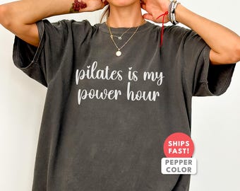以下が含まれることがあります： 「pilates is my power hour」の白い文字がプリントされた濃いグレーのTシャツ。 ゆったりとしたフィット感と半袖です。 右下には赤と白のステッカーが見えます。