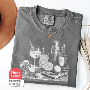 Pode incluir: Uma camiseta cinza Comfort Colors com uma ilustração em preto e branco de ferramentas e ingredientes para fazer coquetéis. Um colar de ouro repousa sobre a camisa. A camisa tem uma etiqueta que diz "S-P-CH". Um adesivo diz "SHIPS FAST! PEPPER COLOR".