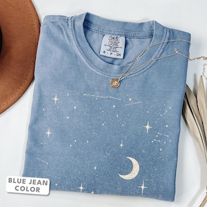 Puede incluir: Una camiseta color azul jean con un estampado gráfico blanco de una luna creciente, varias estrellas y constelaciones. La prenda tiene cuello redondo y una etiqueta de la marca Comfort Colors en el interior. Un collar dorado descansa encima.