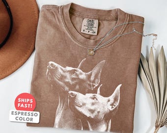 Può includere: T-shirt color espresso con una grafica di due cani con grandi orecchie. La maglietta ha un girocollo e una piccola etichetta. Una collana d'argento con un ciondolo è drappeggiata sulla maglietta. La maglietta è piegata su una superficie bianca.